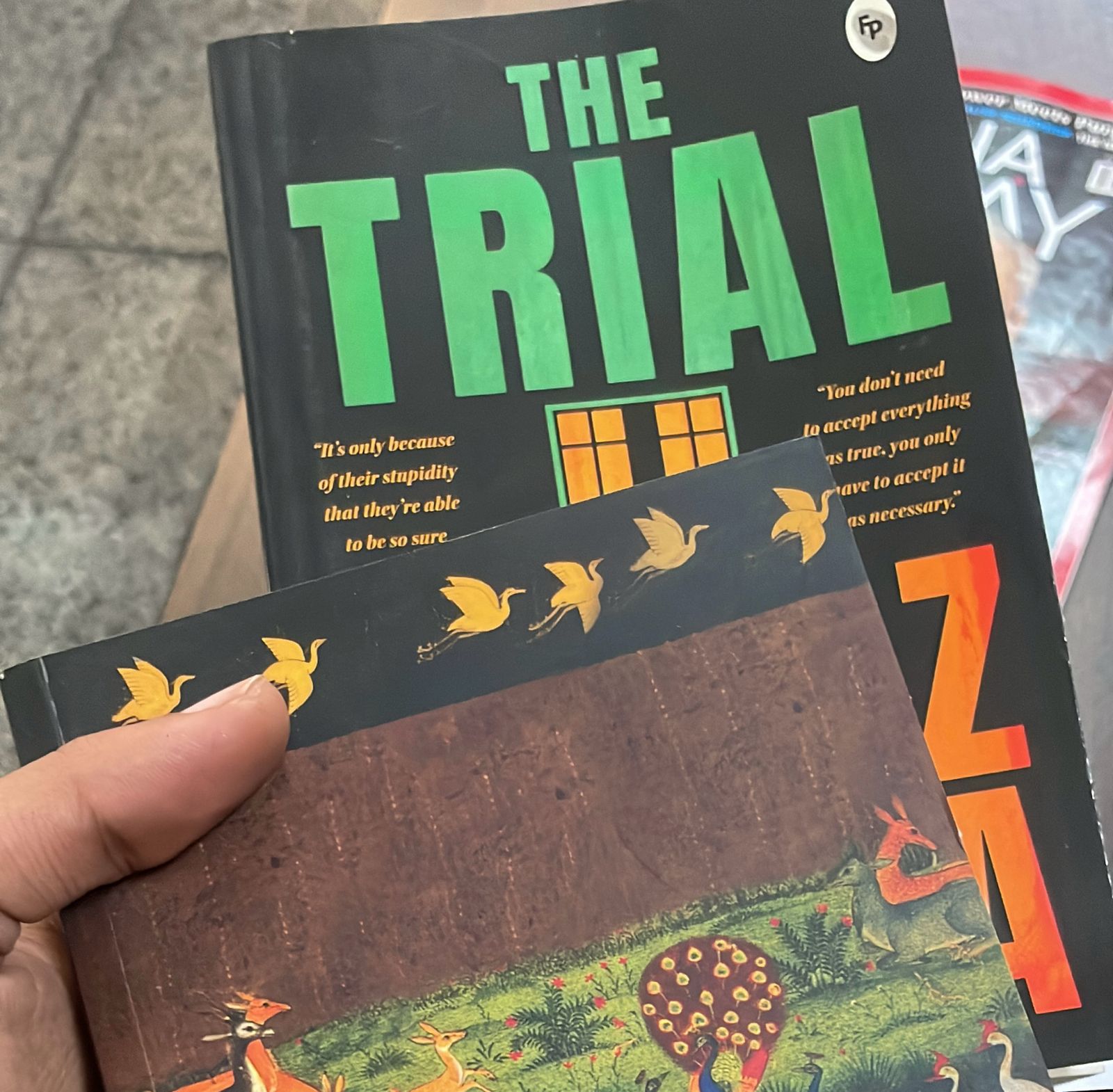 An image of two books held in a hand. The first book is "The Trial" by Frnaz Kafka and the second book is cropped